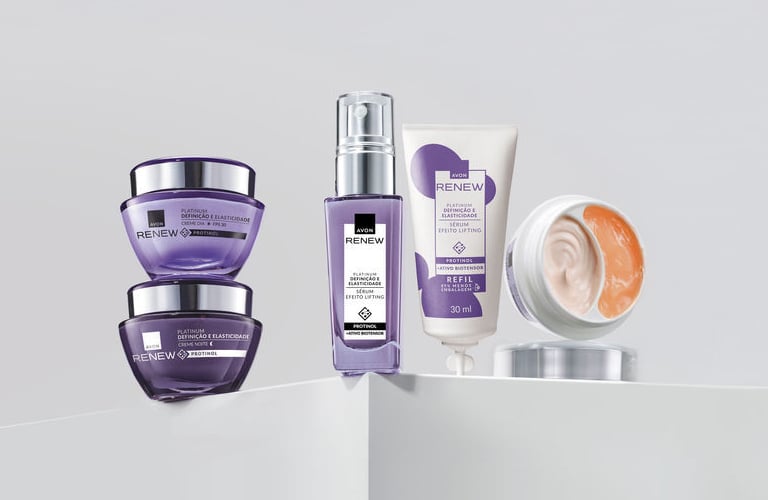 Linha de produtos Platinum Renew da Avon organizados sobre um pedestal branco. À esquerda, dois potes redondos roxos estão empilhados; ao centro, um frasco de sérum roxo e uma bisnaga branca com detalhes em lilás.
