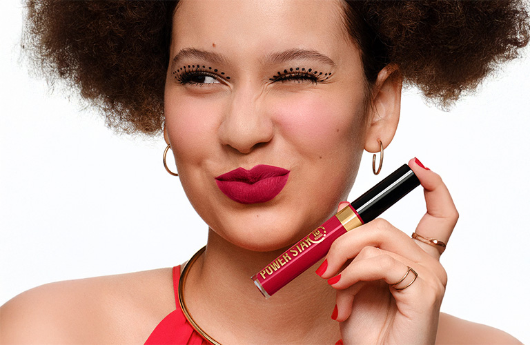 Imagem close-up em rosto de uma mulher branca usando maquiagem de carnaval que contém batom vermelho Power Stay e pontinhos pretos de delineador na pálpebra. Ela pisca um dos olhos e faz um biquinho com a boca.