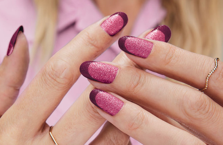 Mãos de uma mulher com unhas coloridas em um estilo de francesinha com uma base em tom rosa e pontas destacadas em um roxo profundo. O visual é complementado por anéis finos e dourado.