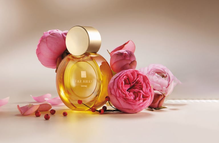 Imagem 02 - Perfume Far Away Shine tem frasco amarelo e tampa dourada e está ao lado de flores rosas, representando a família olfativa floral.