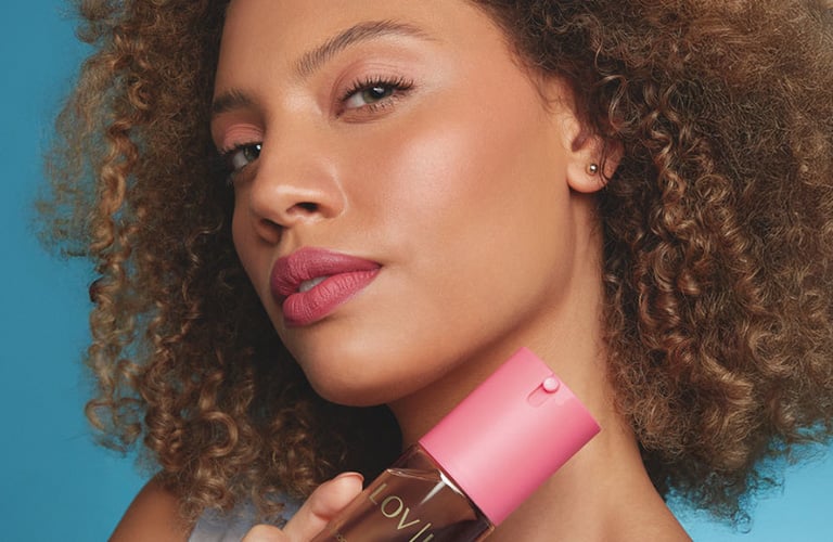 Mulher negra com sorriso discreto e cabelos de curvatura crespa, veste regata branca e segura desodorante corporal em spray da LOV U Avon na cor rosa.