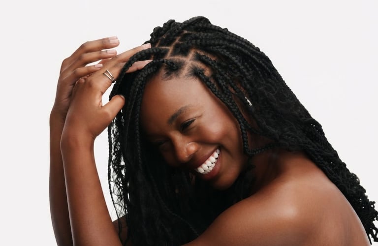 Selo circular com o texto “Uma mulher negra sorri com a cabeça levemente inclinada para baixo, ajustando delicadamente suas tranças com a mão direita. As tranças possuem divisões bem definidas no couro cabeludo e mesclam partes trançadas com pontas e mechas onduladas soltas. Ela veste ombros à mostra e posa contra um fundo branco.