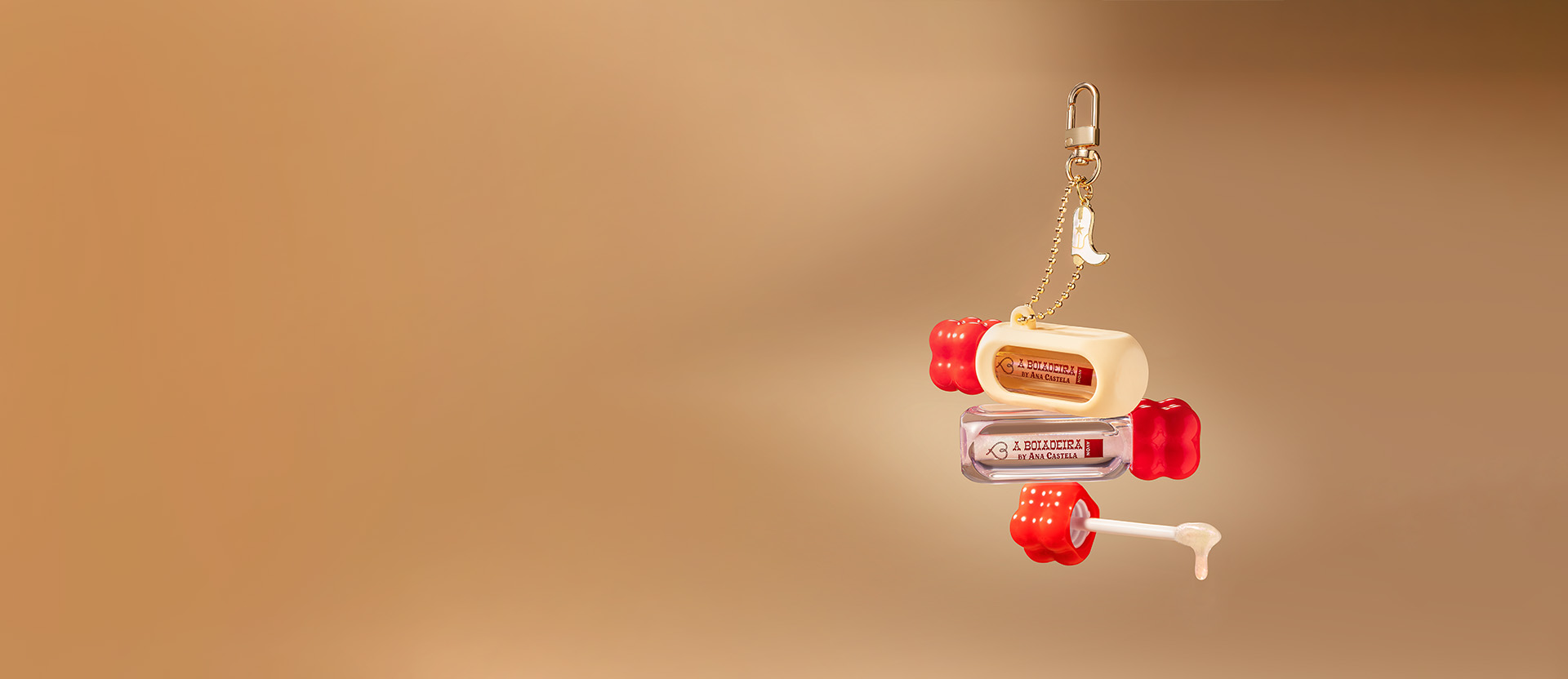 Embalagens com tampa vermelha de Gloss Labial A Boiadeira By Ana Castela, com pingente, capa de silicone e aplicador com textura, em fundo em tons dourados.