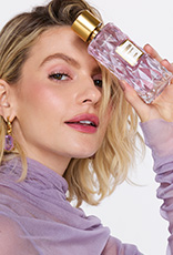 Mulher loira segura frasco da fragrância Charisma Magnetic com tampa dourada e líquido lilás. Ela usa lilás e está em fundo claro.