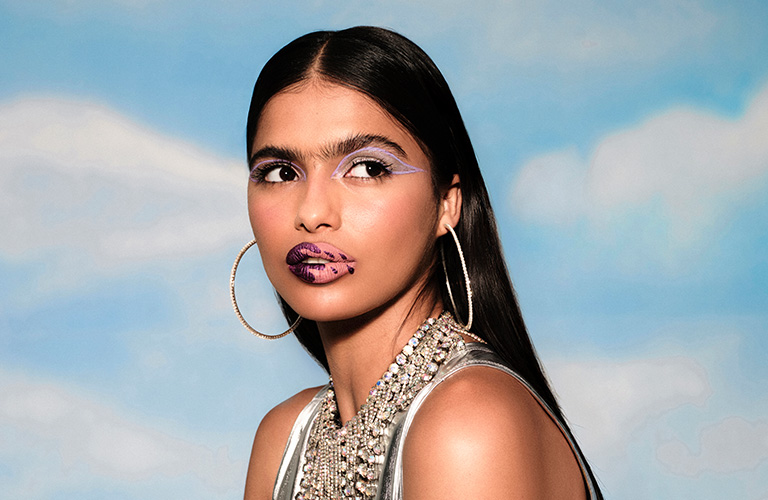 Mulher branca com uma maquiagem de carnaval artística com um delineado gráfico lilás vazado e lábios com desenhos em tons de roxo e lilás. A modelo tem longos cabelos pretos e lisos e usa argolas de strass e um top prata metalizado.