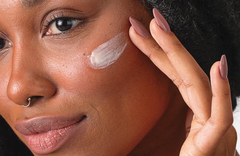 Mulher negra com um sorriso suave enquanto espalha um creme branco na maçã do rosto com as pontas dos dedos. Ela tem os cabelos escuros trançados, unhas pintadas em tom rosado e olha para a lateral da imagem.