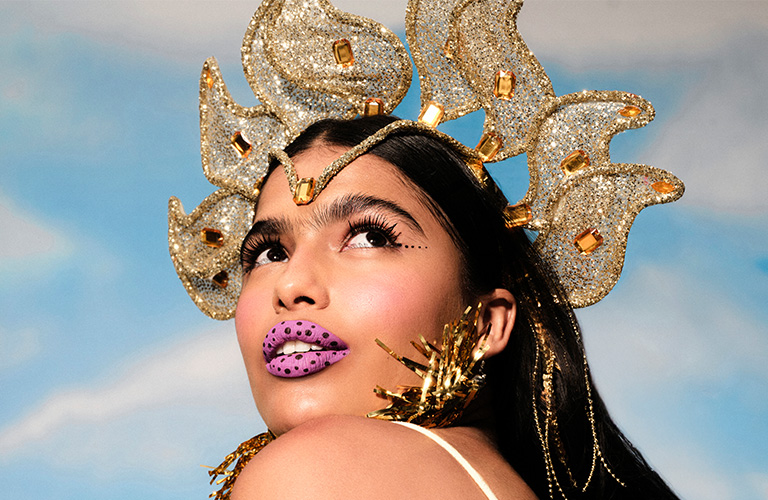 Mulher branca com longos cabelos pretos olha para cima, exibindo uma maquiagem de carnaval artística. O grande destaque são os lábios em tom lilás vibrante com bolinhas pretas sobrepostas. Ela usa uma coroa dourada com brilhos e pedrarias. O fundo da imagem é um céu azul claro com nuvens brancas.