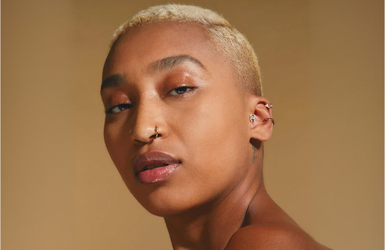 Mulher negra com cabelo curto, raspado nas laterais e descolorido em tom loiro-claro. Ela posa olhando diretamente para a frente com os lábios levemente entreabertos. Sua maquiagem em estilo natural destaca as pálpebras com um acabamento brilhante de efeito “glossy” em tom bronze ou marrom-claro.