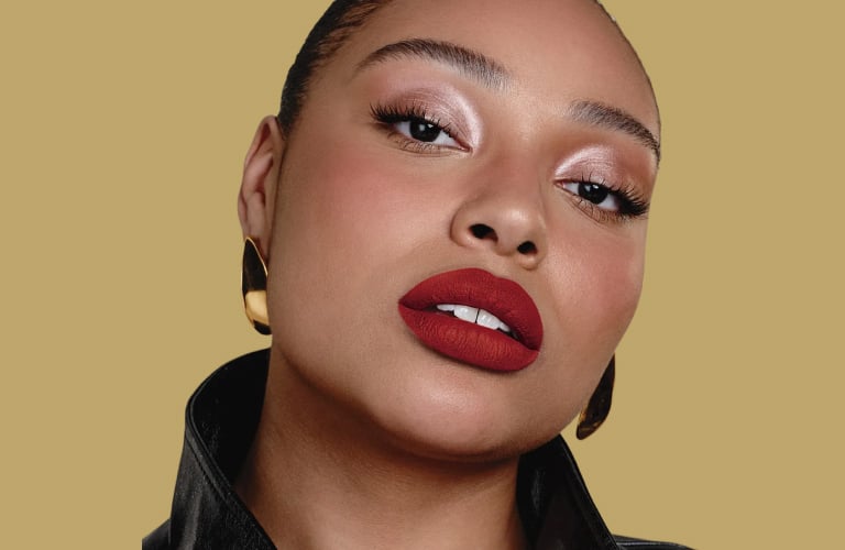 Mulher com pele oliva olha diretamente para foto e está maquiada com batom vermelho escuro e sombra brilhosa em tons prata e marrom.