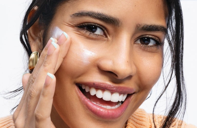 Mulher sorridente de cabelos castanhos longos amarrados, veste camisa regata laranja e aplica protetor solar Renew no rosto.