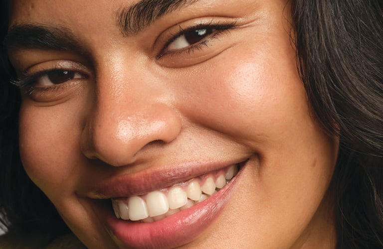 Mulher de pele parda, com sobrancelhas marcantes e cabelos pretos. Ela exibe um sorriso largo, olhando diretamente para a câmera.