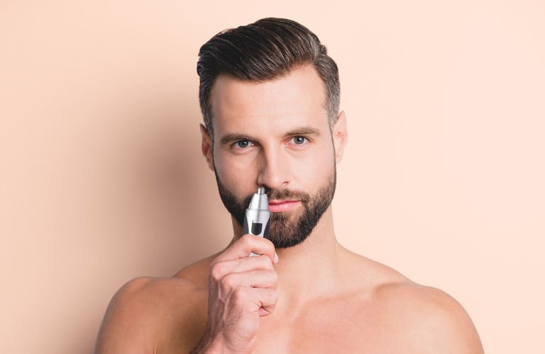 Homem de pele clara, com barba e cabelos castanhos curtos, sem camisa contra um fundo bege. Ele segura um aparador de pelos pequeno próximo ao nariz.