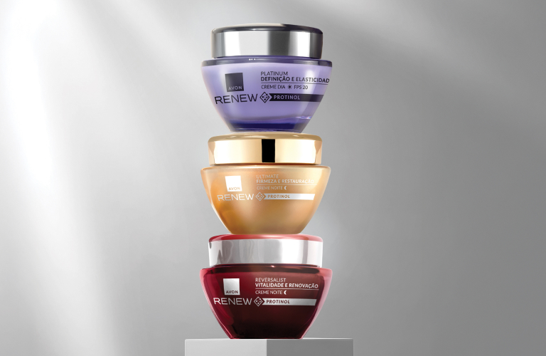 Três potes de cremes antissinais da Renew da marca Avon empilhados verticalmente sobre um pedestal branco. O pote da base é rosa escuro, o do meio é bege claro e o do topo é lilás, todos com tampas prateadas e rótulos arredondados.