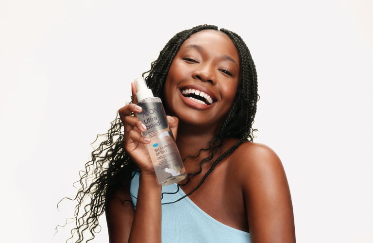Selo circular com o texto “Uma mulher negra sorri segurando o frasco transparente do Shampoo Spray Avon Ultra Braids próximo ao rosto, destacando suas tranças no cabelo. Ela veste uma blusa azul-clara de alça única sobre fundo branco. As tranças no cabelo são finas, escuras e terminam em mechas onduladas soltas que acompanham o movimento do seu corpo.