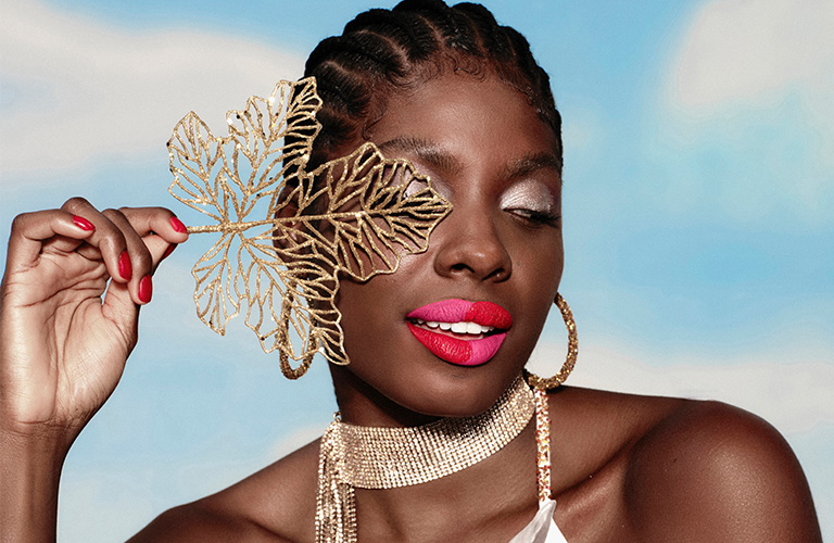 Mulher negra com cabelos trançados exibindo uma maquiagem de carnaval no estilo color block. Seus lábios estão divididos artisticamente em quatro quadrantes, intercalando tons de rosa vibrante e vermelho intenso. Ela posa segurando uma folha dourada com glitter sobre um dos olhos, que está decorado com sombra prata cintilante.