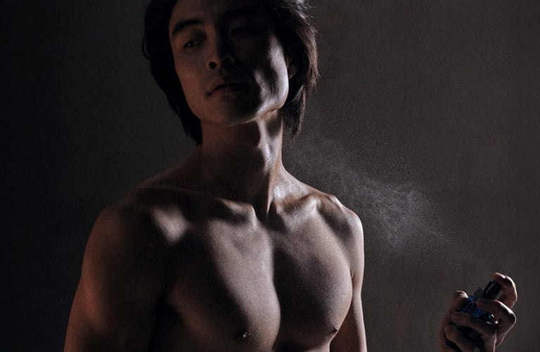 Homem com traços nipônicos (olhos amendoados, rosto oval e nariz fino) e cabelos pretos lisos está sem camisa, enquanto borrifa perfume masculino no peitoral.