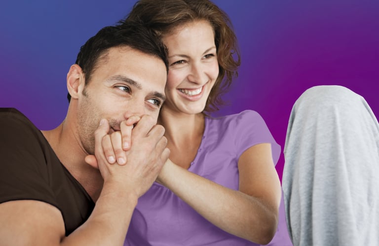 Um casal sorridente e abraçado em frente a um fundo em degradê azul e roxo. O homem veste camiseta escura e beija carinhosamente as mãos da mulher, que está de camiseta roxa e sorri olhando para o lado.