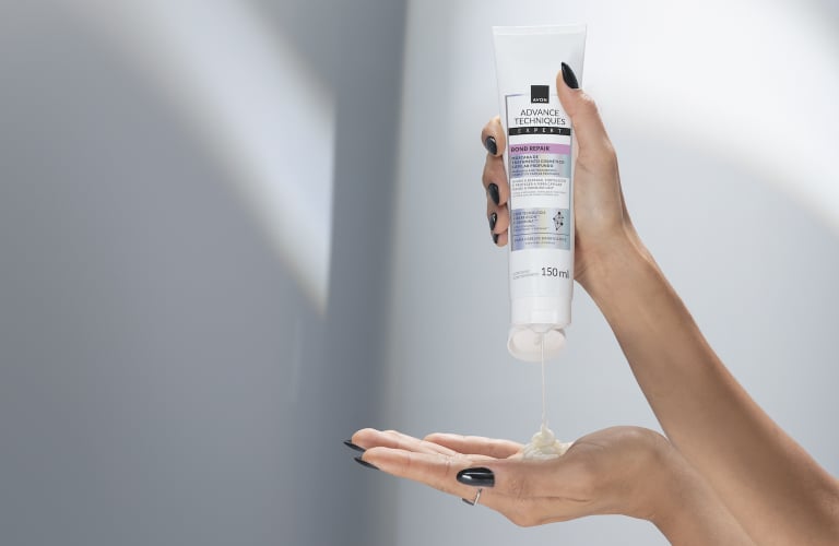 Mãos de uma pessoa de pele clara com unhas pintadas de preto segura uma bisnaga branca de máscara capilar da linha Bond Hair da Avon, pressionando-a para despejar uma porção de creme na palma da mão direita.
