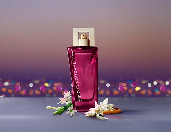 Fragrância Avon Attraction Closer para Ela em bancada cinza com ingredientes, como cítricos e flores, em fundo de entardecer de uma cidade iluminada.
