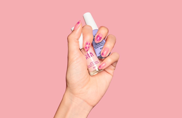 Mão segurando dois vidros de esmalte. O primeiro é um top coat e o outro uma base de frasco roxo. As unhas estão pintadas de rosa com detalhes decorativos.
