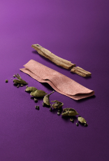 Amostras de cardamomo, couro e sândalo, ingredientes nobres da fragrância Black Essential Charm, aparecem em destaque no centro da tela sobre fundo violeta.