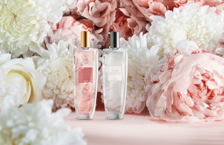 Um frasco de perfume Pur Blanca Avon e uma loção hidratante da mesma linha posicionados ao centro, cercados por um arranjo de flores em tons de branco e rosa-claro, sobre um fundo rosa suave.