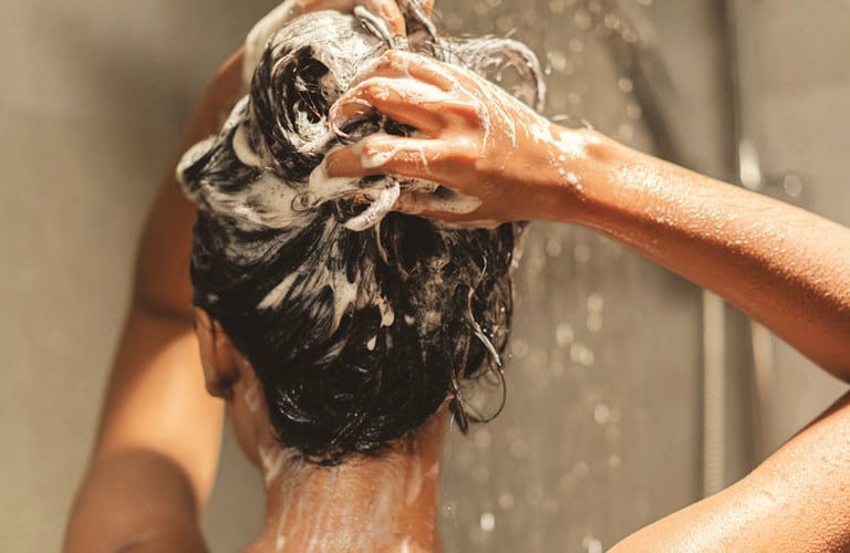 Mulher de costas durante o banho premium, com os braços erguidos enquanto ensaboa os cabelos escuros. Há bastante espuma branca acumulada no topo da cabeça e sabão escorrendo pelas costas.
