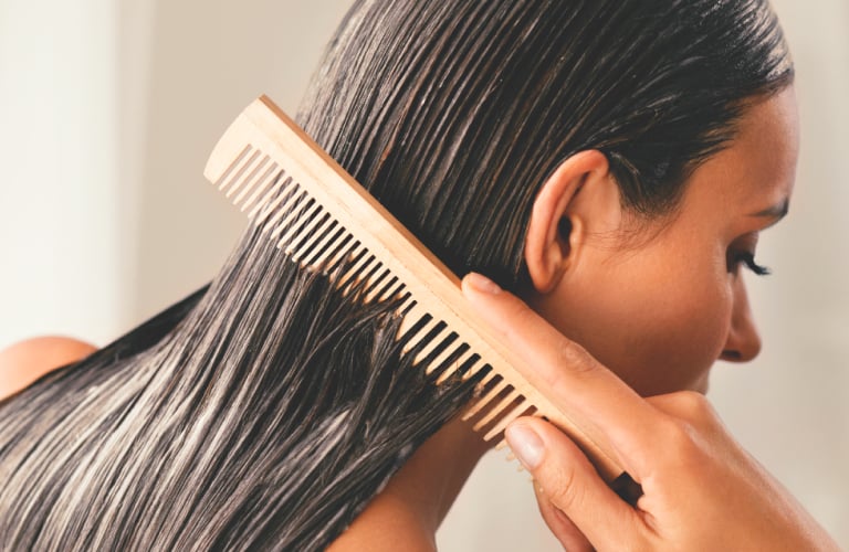 Mulher de costas penteia o cabelo liso e úmido com pente de madeira, aplicando Shampoo Limpeza Profunda da Avon.