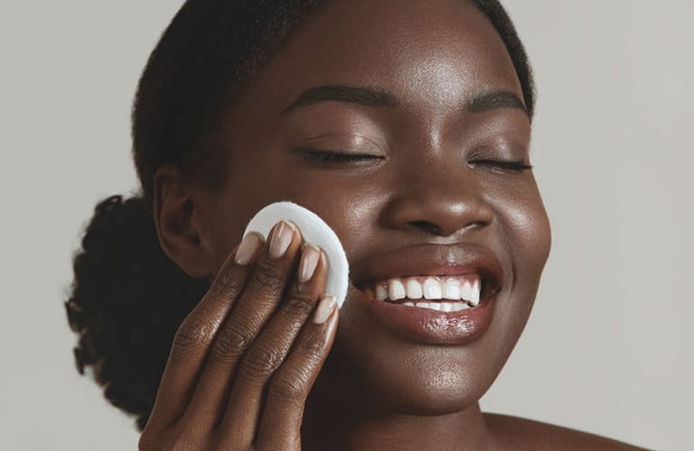 Mulher negra de pele retinta e cabelos presos, sorrindo com os olhos fechados. Ela está encostando um disco de algodão branco no rosto, em um gesto de cuidado com a pele pós-depilação facial.