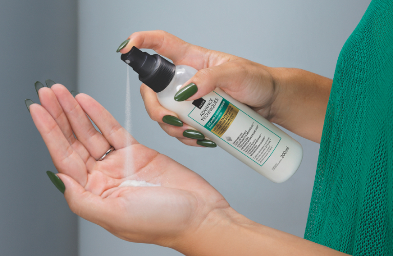 Mulher de blusa verde borrifando a loção de tratamento day after em spray da Avon na palma da mão para aplicar no cabelo.