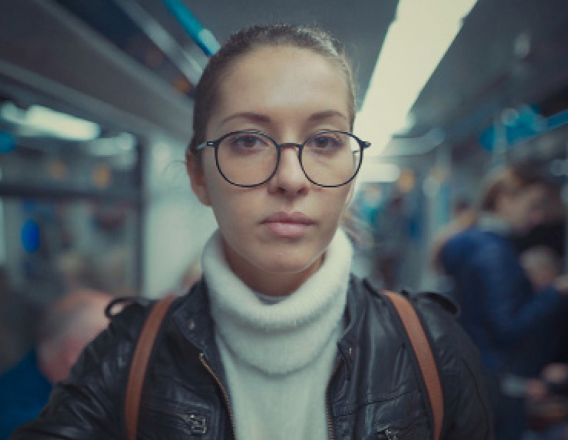 Foto de mulher branca de cabelos presos, com blusa de gola alta branca e jaqueta de couro preta olhando para a câmera. O fundo é a imagem desfocada do interior de um vagão de metrô.
