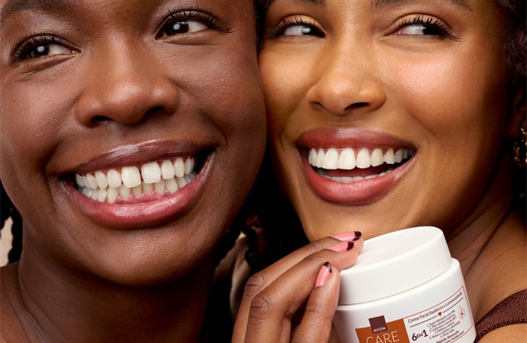 Duas mulheres sorridentes, uma ao lado da outra. À esquerda, uma mulher de pele retinta e cabelos com tranças nagô. À direita, uma mulher de pele parda com cabelos cacheados. Elas seguram um pote branco do creme facial da Avon Care de Luminosidade e Radiância.