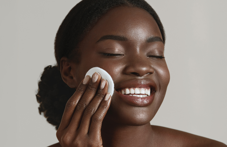 Mulher negra sorrindo com os olhos fechados enquanto passa um disco de algodão branco no rosto radiante. Ela está com os cabelos escuros presos em um coque baixo e os ombros à mostra, contra um fundo neutro em tom claro.
