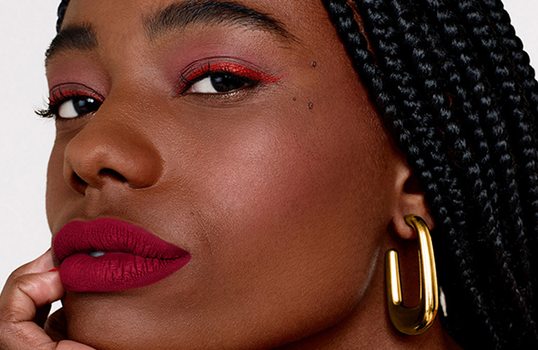 Mulher negra de tranças usando batom matte magenta, delineado gatinho vermelho e sombras rosadas. Ela posa com a mão no queixo e usa brincos dourados.