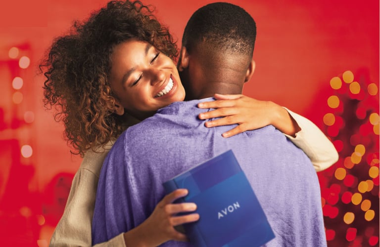 Mulher de cabelos com curvatura 4A-4C está sorridente, segurando um presente de natal da Avon, enquanto abraça um homem que está de costa (seu rosto não aparece na foto).