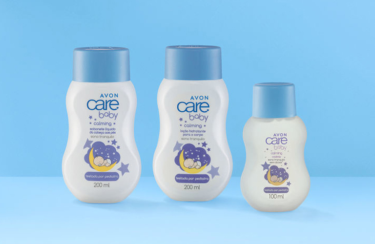 Três frascos brancos com detalhes roxos da linha Avon Care Baby sobre fundo azul-bebê para o kit de mãe de primeira viagem.