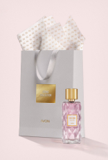 Frasco em vidro facetado e tampa dourada de Charisma Magnetic à frente de sacola de presente cinza com texto Iconic Collection Avon, e papel de seda branco. Fundo claro.
