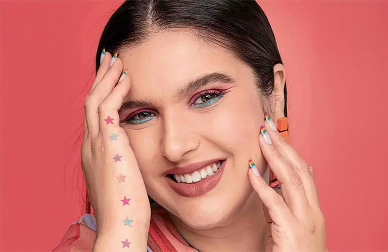 Mulher branca sorridente exibindo uma maquiagem de carnaval colorida. Ela destaca o olhar com sombra rosa esfumada e um delineado azul turquesa rente aos cílios inferiores. O visual é complementado por unhas estilo francesinha com pontas arco-íris e uma fileira de pequenas estrelas coloridas aplicadas na lateral da mão.