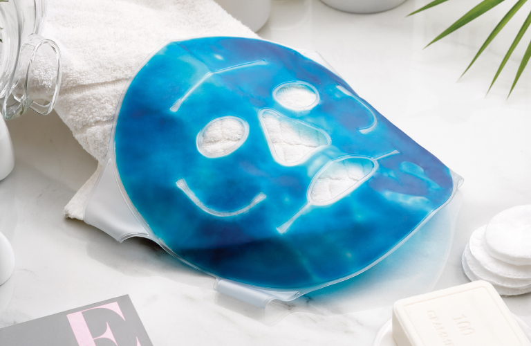 Máscara facial de gel azul disposta em uma superfície clara de mármore. Ao fundo, há velas aromáticas, potes com inscrições, folhas de palmeira e, ao lado, um sabonete em barra e toalhas dobradas para um momento de cuidados com a pele.