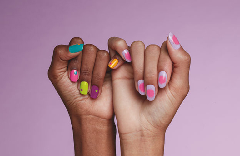 Dois braços erguidos contra um fundo lilás suave, com as mãos se tocando para exibir unhas coloridas com cores diferentes entre elas, como verde-água, amarelo e rosa com detalhes brancos.