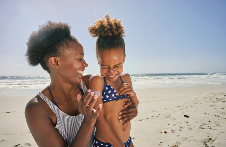 Mulher negra sorridente aplicando protetor solar no rosto e nos braços em uma menina de biquíni azul de bolinhas brancas em uma praia ensolarada, com o mar e a areia ao fundo.