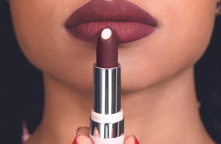 Close-up nos lábios de uma pessoa usando batom matte em tom de vinho. Ela segura o bastão do batom centralizado à frente da boca, destacando o acabamento opaco.