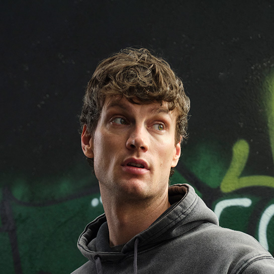 Homem de cabelos curtos usando moletom cinza com capuz, em fundo com grafites em tons de verde.