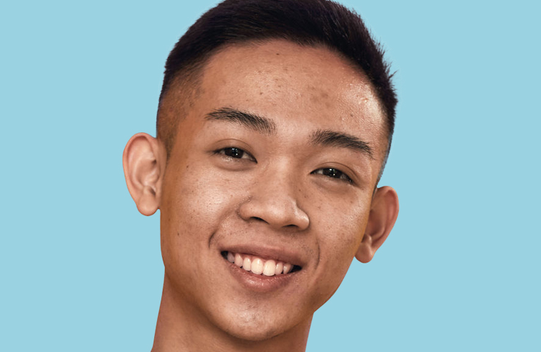 Adolescente homem de ascendência asiática sorrindo. Ele tem cabelos pretos curtos, raspados nas laterais, e posa contra um fundo azul-claro sólido.