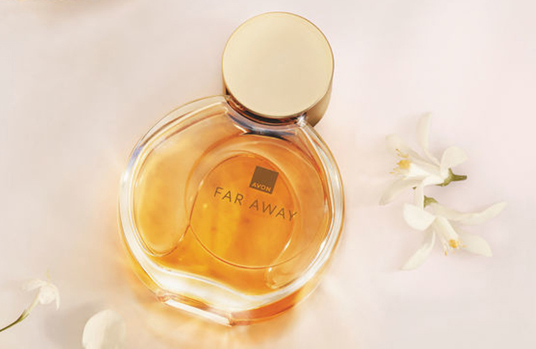 Frasco de perfume Far Away da Avon, com líquido em tom âmbar e tampa dourada, centralizado sobre um fundo suave em tons creme e decorado com pétalas soltas de um tipo de jasmim.