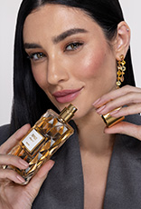 Mulher branca de cabelos lisos segura frasco da fragrância Topaze Elegant com tampa dourada e líquido pêssego. Ela usa roupa cinza e está em fundo claro.