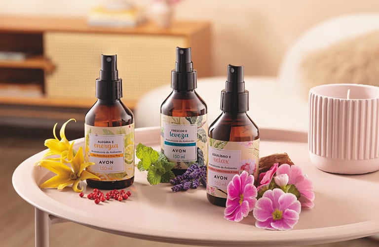 Três frascos de aromatizador de ambiente sobre uma bandeja rosa com flores naturais e uma vela perfumada, em um ambiente de casa.