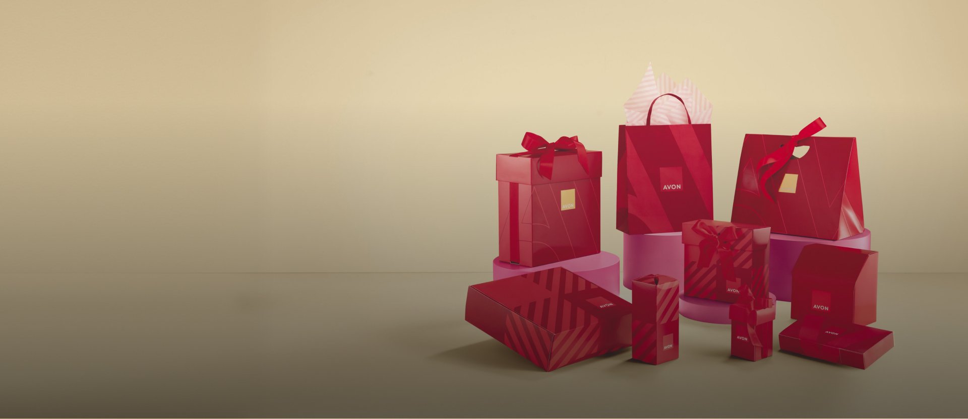 Diferentes embalagens de presente vermelhas da Avon, incluindo caixas e sacolas, com o logo Avon em destaque. Elas estão dispostas sobre plataformas rosa. Fundo bege.