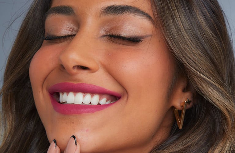 Mulher sorridente de cabelos ondulados castanhos usa uma maquiagem de noiva de batom rosa, sombra cintilante e pele iluminada.