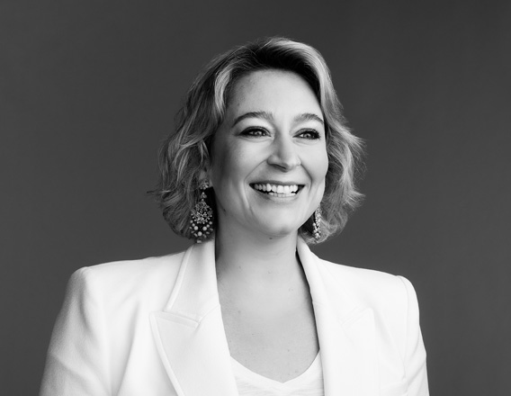 Foto em preto e branco de mulher sorrindo com cabelos loiros médios, usando roupa branca e brincos. 
